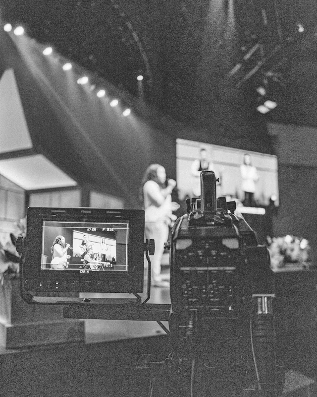 A video camera recording a stage, its viewfinder in the foreground